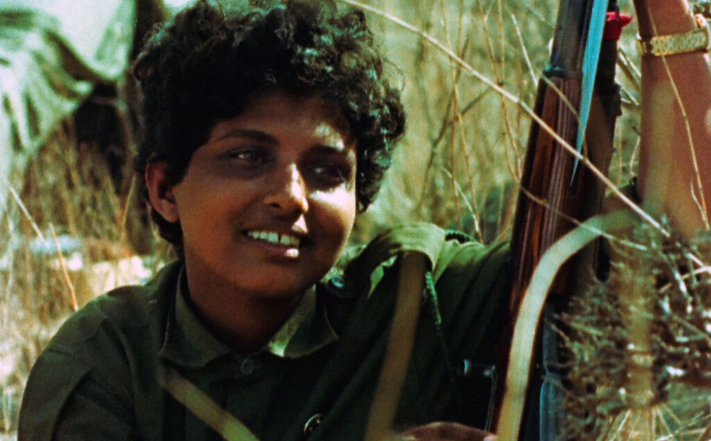 Closeup of young woman with short dark hair sitting and smiling in the grass, holding her gun upright