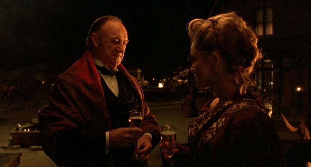John Herod (Gene Hackman) in a suit and red dinner jacket, eyes Ellen “The Lady” McKenzie (Sharon Stone) in an ornate maroon dress. It’s night. The two are standing outside in front of Herod’s house. Both are holding drinks and sizing each other up.