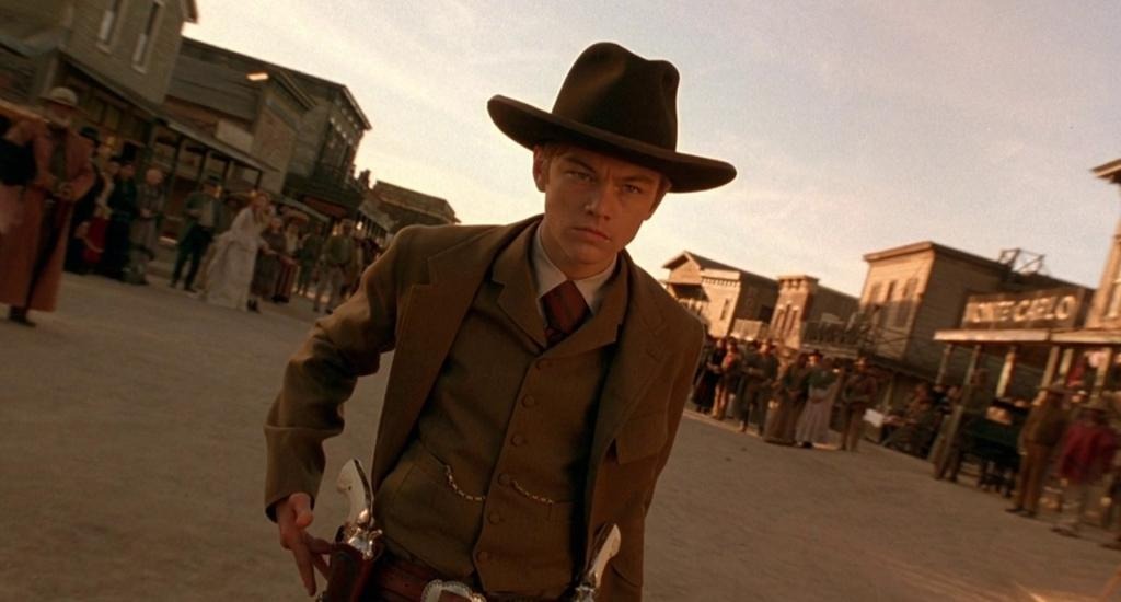 Fee “The Kid” Herod (Leonardo DiCaprio) stares sternly at an enemy offscreen. He’s dressed in a dapper brown suit and cowboy hat, at the center of town square, fingering his gun in its holster, surrounded by onlookers.