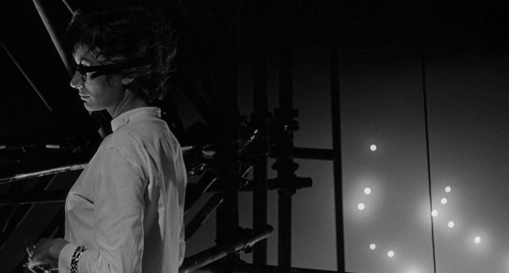 A stylish woman in designer glass stands on scaffolding on a movie set.