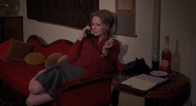 woman reclining on a sofa, smoking a cigarette and talking on the phone. She wears a maroon cardigan and pleated grey skirt. 