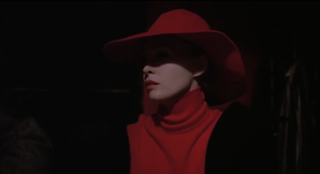 Medium shot of woman in a red cowl-necked dress and red fedora, her lipstick the same shade of red. She is surrounded by the darkness of a bar. 