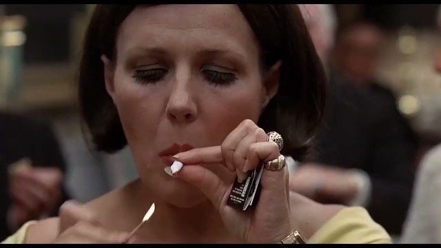 A close-up of a woman with a dark-brown bob lighting a joint with a match, eyes closed and lips pursed. Other figures are in soft focus behind her, and in front of her, the lit match is approaching the end of the joint, which she holds between her thumb and forefinger, the rest of her fingers bejeweled and clasping a black-and-white matchbook. She's wearing a gold watch, and while her clothes are mostly not visible due to the proximity of the shot, her top appears to be a wide-necked canary yellow.