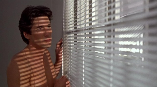 Richard Gere stands to the left of the frame, shown from his mid-chest up. He has the shadows of Venetian blinds going across his bare chest and face. He has brown hair and is gazing out the window. Filling two thirds of the right side of the frame are the Venetian blinds, at an angle and getting closer to the viewer in the foreground. Light is coming through the blinds and his right hand is resting against them at the bottom while his left hand is held up and resting against the wall to the left of the blinds. He is shirtless and gazing out the window.