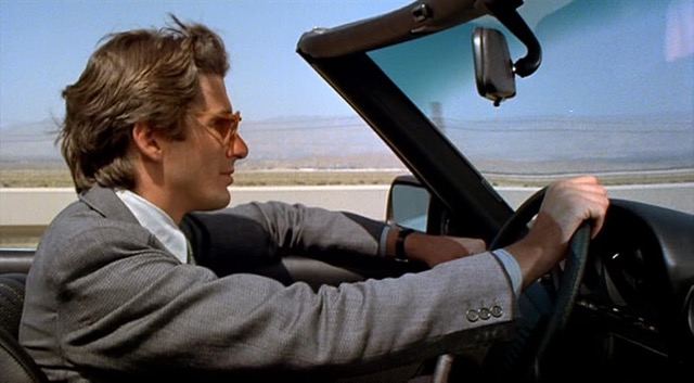 Side view of Richard Gere driving a convertible and wearing a nice gray suit with a white collar. His brown hair stands up at his forehead and is swept back ever so slightly and he is wearing sunglasses, with his arms at the steering wheel. He is facing towards the right of the frame in profile. In the distance on his left side and behind him you see a white wall and some kind of field beyond it that fades into the background and into the blue sky above it.

