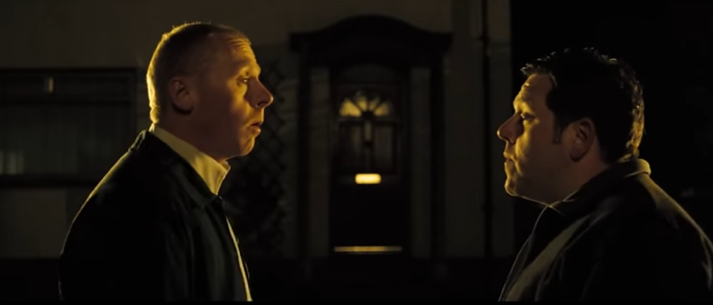 White male with short blonde hair looks at a shorter man with black hair in front of a door. Both have on black jackets.
