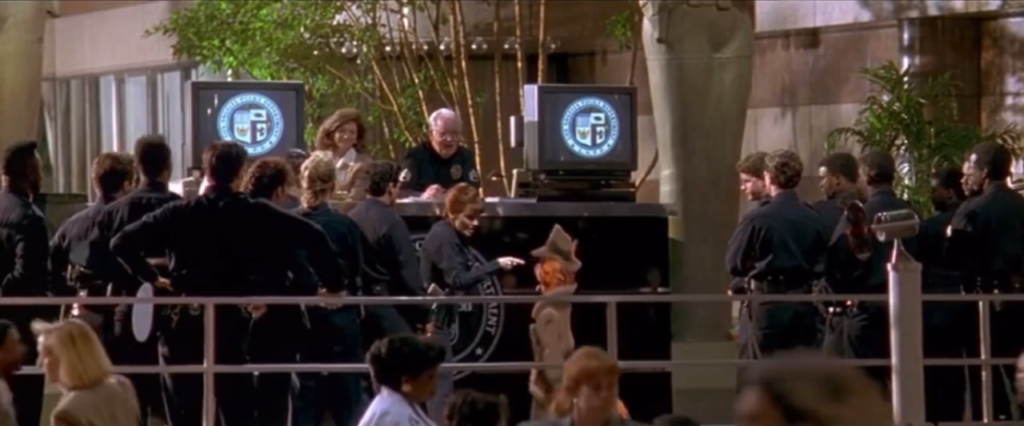 A woman with red hair in black police uniform points at an animated tall orange anthropomorphic cat in a tan trench coat in front of a desk with another police officer. 2 lines of police officers are behind them.