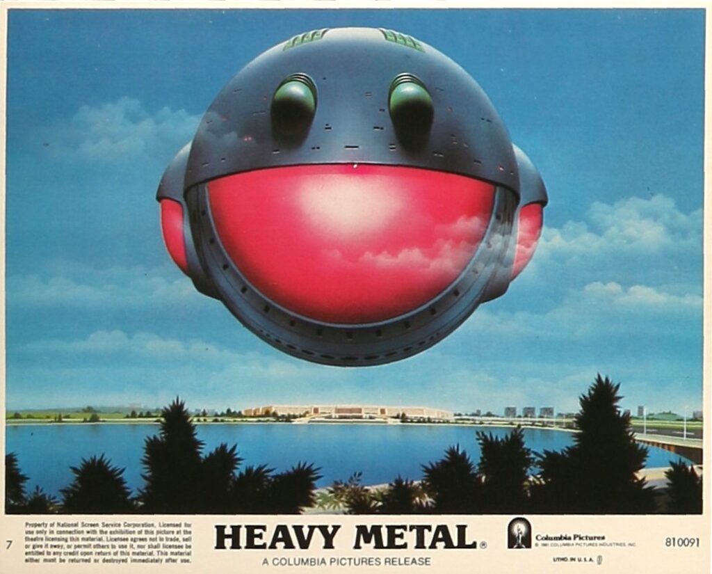 Publicity still from Heavy Metal showing a large, spherical spaceship hovering over a calm body of water near a modern complex of low buildings. The craft is metallic gray with two green protrusions on top and a wide, glowing red front window. Dark tree silhouettes frame the foreground, and the sky is bright blue with scattered clouds. Text along the bottom reads “HEAVY METAL – A Columbia Pictures Release.”