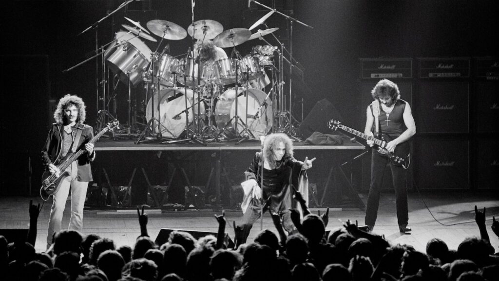 Black-and-white concert photo of a heavy metal band performing onstage. The singer stands at center, leaning forward dramatically toward the audience with one hand extended. A guitarist and bassist flank him on either side, while a drummer plays on a raised platform behind them, surrounded by a large drum kit and cymbals. Tall amplifier stacks line the back of the stage. In the foreground, a packed crowd raises hands in the air, some making the “devil horns” gesture.