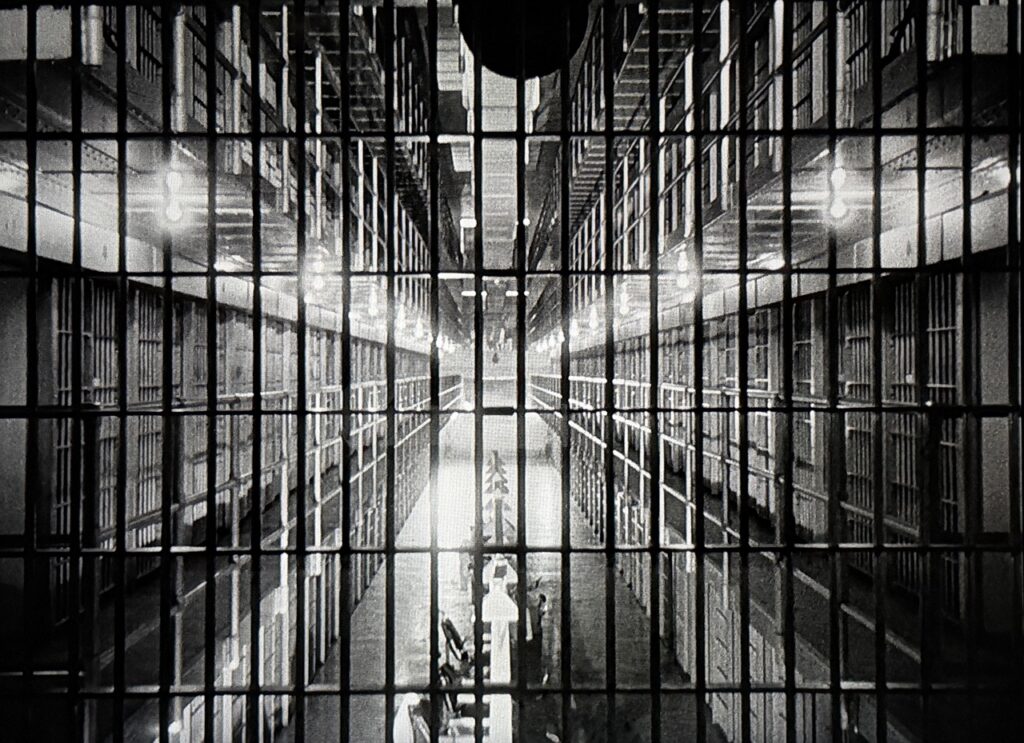 A semi-abstract image of prison bars filling the frame and, behind them, the lights of a prison cell block.