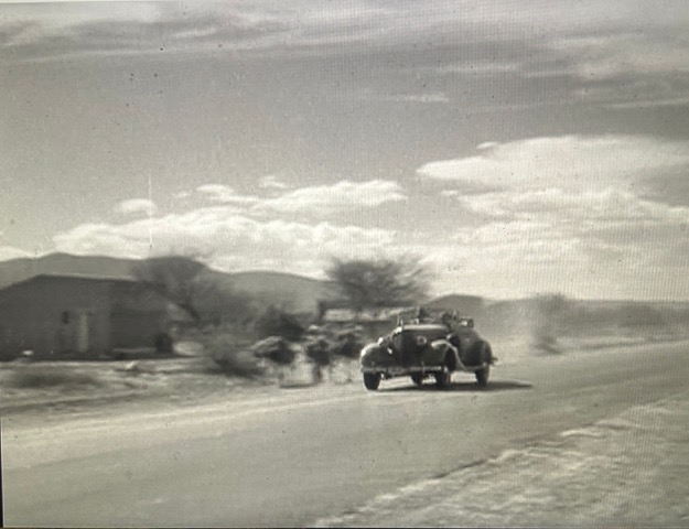 A black and white image of a car driving down the road with two passengers inside. Dust rises from the road, and the trees appear to be dead.