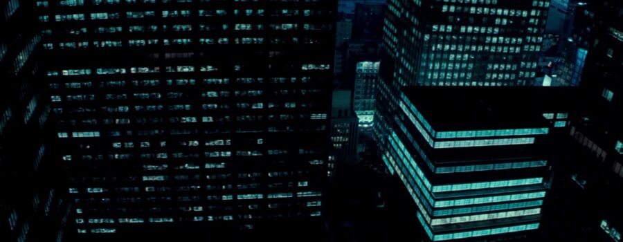 A cold, lit up jumble of New York skyscrapers at night, shot from overhead, just close enough to get a hint of the activity that populates the office behind each window.