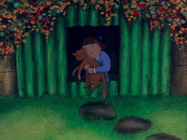 Picture of a girl in a bunker with green grass and flowers over the top of the bunker. She is hugging a cat that has approached her and she is smiling, as is the cat.