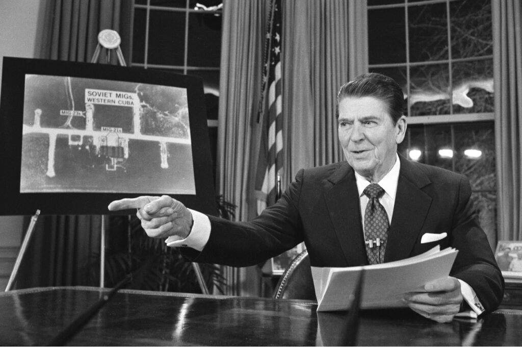 Black and White picture of Ronald Reagan in the White House. He is pointing at something out of frame. He is holding a paper script and behind him is a chart that says “Soviet MiGs-Western Cuba” and it appears to be a satellite photo.