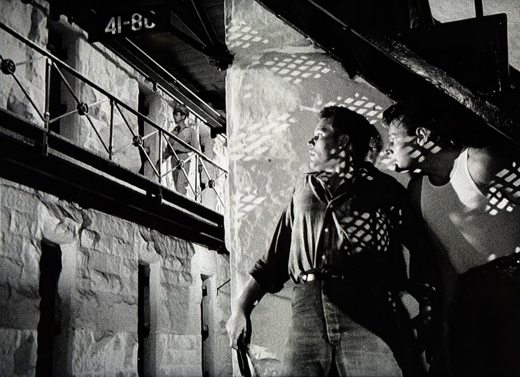 In the foreground, two men in prison uniforms hug a wall. In the background, a prison guard patrols along the uppier tier of a cell block.