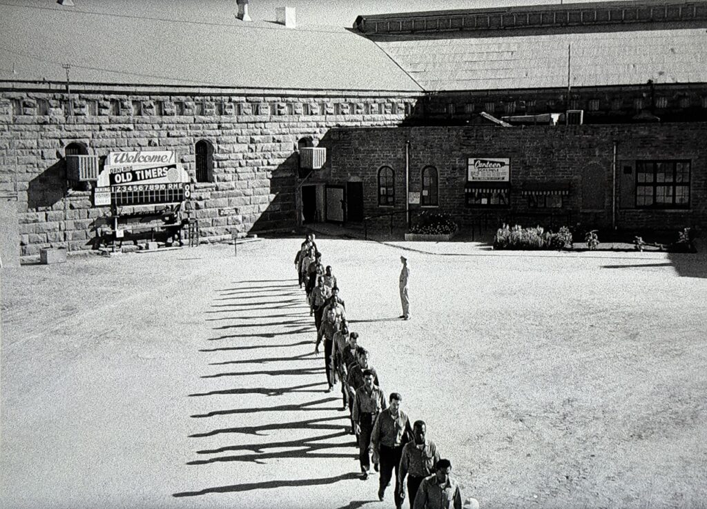 Wide-angle image of a prison exercise yard. Prisoners march single-file from the top to the bottom of the frame.