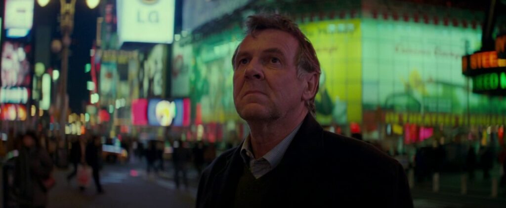 Arthur Edens (Tom Wilkinson) stands in the middle of a lit up Times Square and looks seriously off camera. 