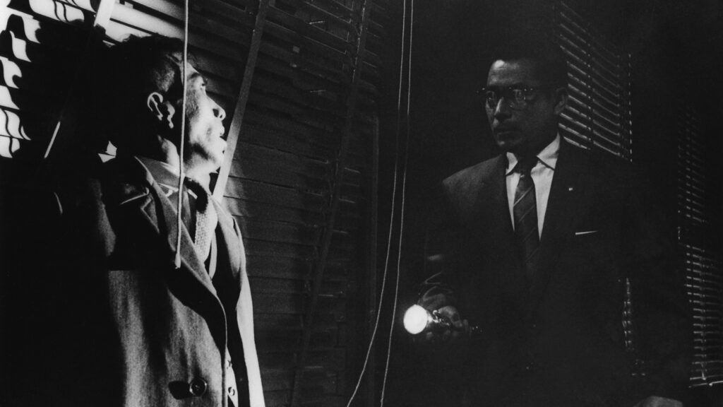 Nishi shining a flashlight on a scared Shirai in the dark.