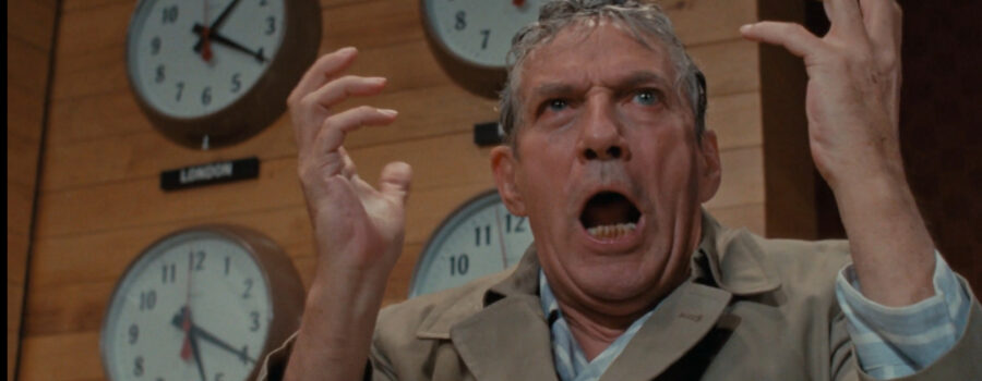 Peter Finch as Howard Beale standing in front of a bank of clocks, sweaty and angry, his hands in the air, yelling.