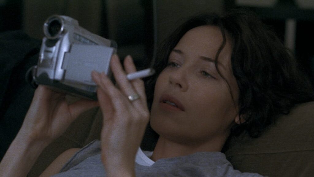 Diane (Connie Nielsen), a woman with short brown hair, reclines on a couch and looks at the viewfinder of a digital camcorder. Her mouth is slightly open in an inscrutable expression of engrossment. She holds a cigarette between her fingers.