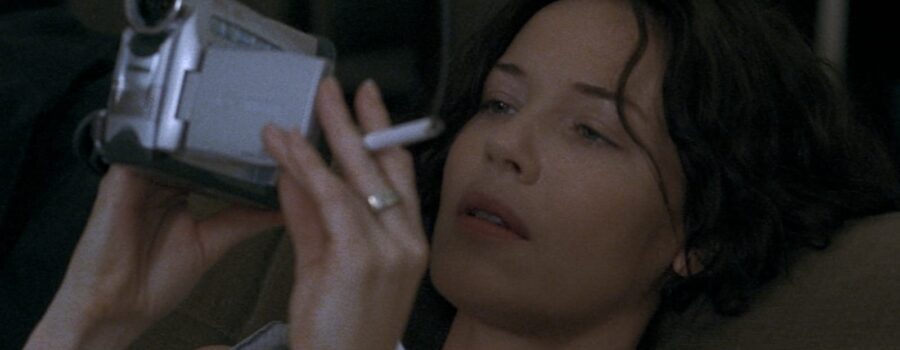 Diane (Connie Nielsen), a woman with short brown hair, reclines on a couch and looks at the viewfinder of a digital camcorder. Her mouth is slightly open in an inscrutable expression of engrossment. She holds a cigarette between her fingers.