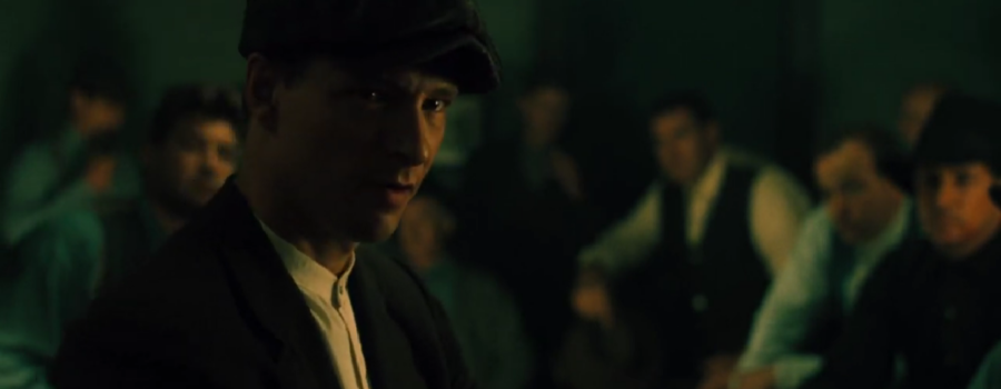 A white man with a stern expression on his face wearing a snap-brim cap and 1920s period clothing delivers a speech at a meeting as a group of men surrounds him listening intently to the speech.