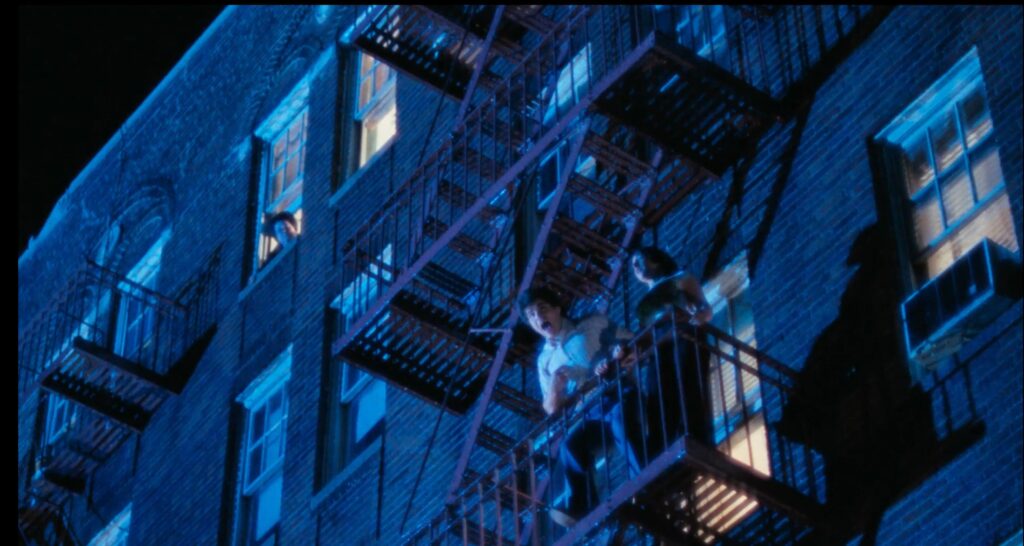 A couple stands on a fire escape in an apartment building in the rain. Lightning is flashing, illuminating them. The man is yelling. A woman in the top left corner of the frame is looking down at them.