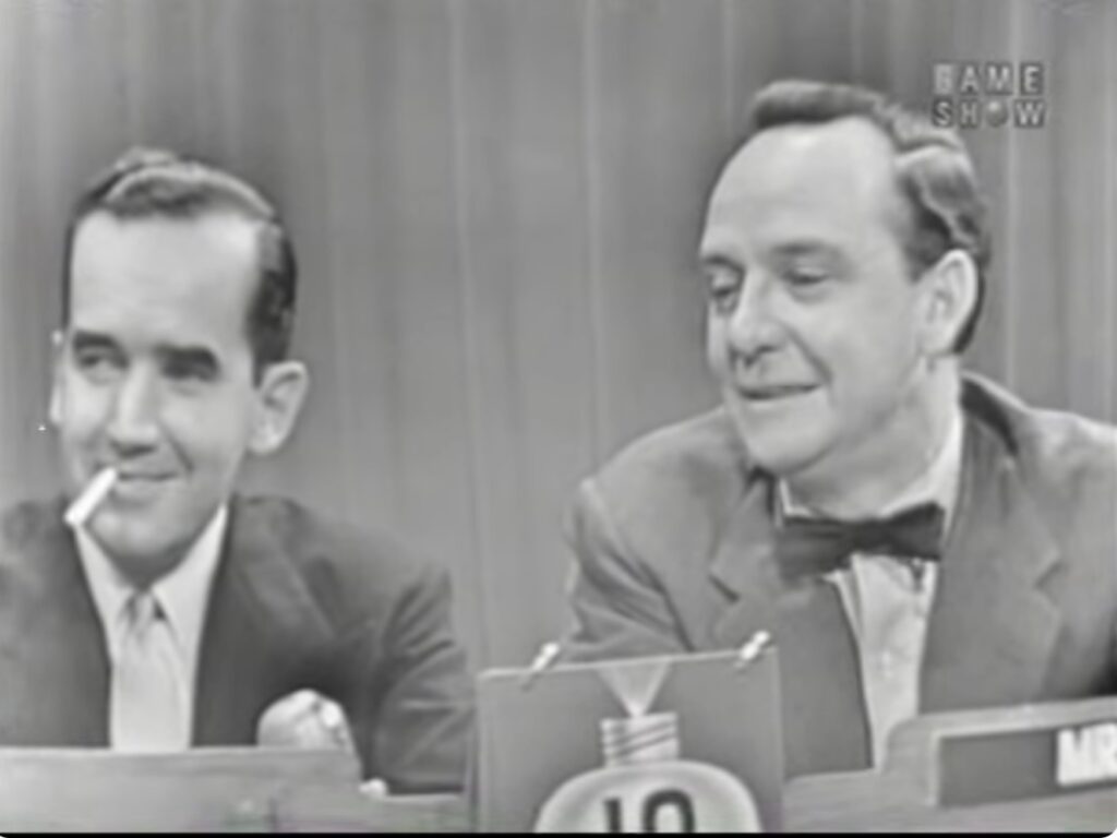Black and white screenshot of Edward R Murrow, smiling, sitting next to the host of the game show What’s My Line, John Daly