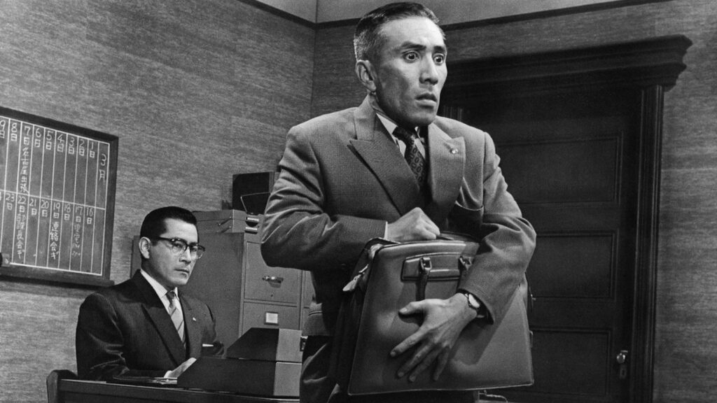 A petrified Shirai (Kō Nishimura) holding his briefcase with Nishi (Toshiro Mifune) observing from his desk.