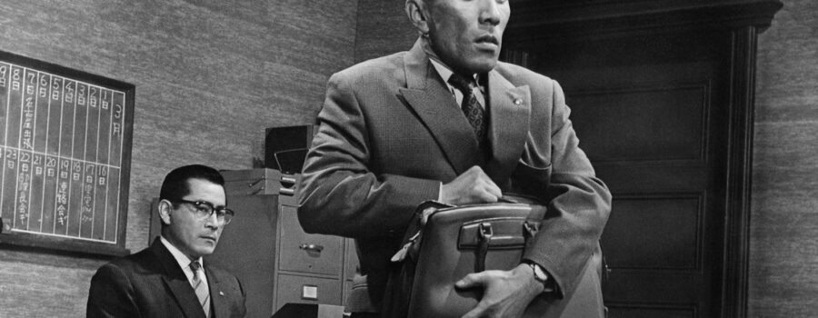 A petrified Shirai (Kō Nishimura) holding his briefcase with Nishi (Toshiro Mifune) observing from his desk.