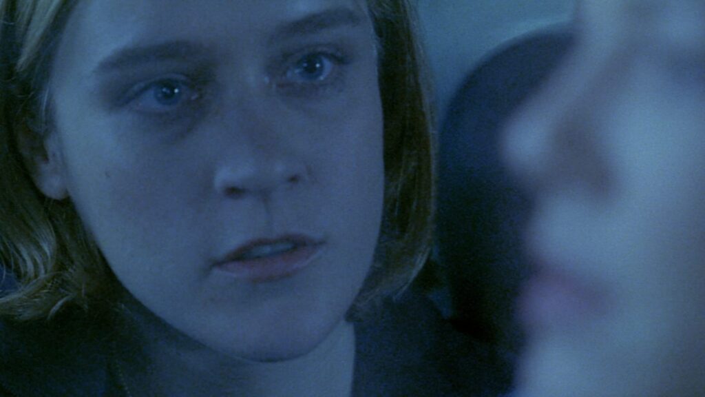 Elise (Chloë Sevigny), a blonde-haired woman with blue eyes, looks at a person in the foreground, who is blurred out of focus. Elise has a focused look on her face and her lips are pursed, as if mid-speech.