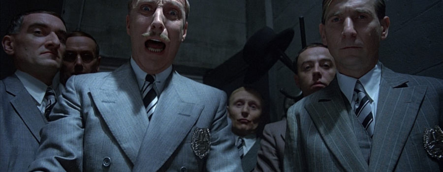 A group of six looming, smirking white men in light grey, pinstripe suits looking into camera. The central man is agape in frustrated anguish.