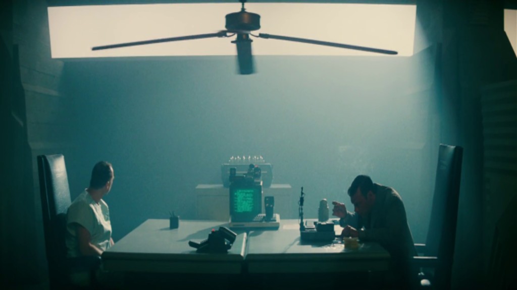 Leon (Brion James) sits across Holden (Morgan Paull) in an interrogation room. The Voight-Kampff testing machine rests on the long table between them.