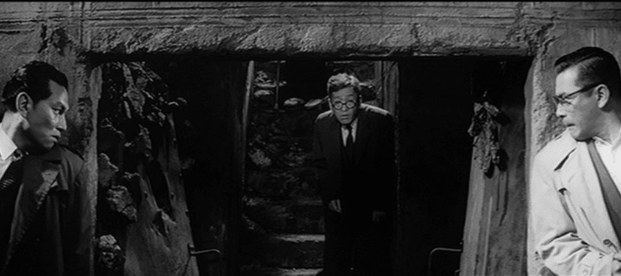 Itakura (Takeshi Katō), left, and Nishi, right, hide around the corners as an unsuspecting Wada (Kamatari Fujiwara) returns to their hideout.