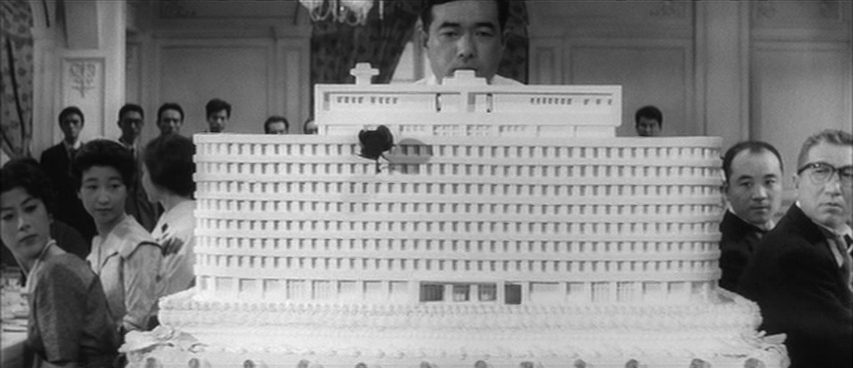 Cake shaped like the Public Corporation building being rolled out with the guests looking it over.