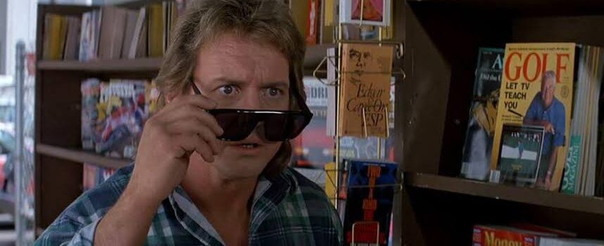 John Nada (Roddy Piper), a white adult man with a dark blonde mullet hair cut, wearing a plaid short, is looking up from his dark sunglasses. He is standing in front of a news stand.