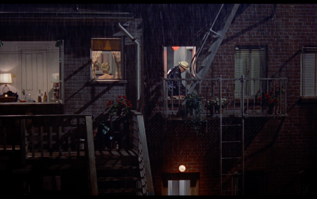 An exterior shot of a brick apartment building. Through one window there’s a woman with blonde hair, and through an adjacent window there’s a man in a cream hat and black coat.>
