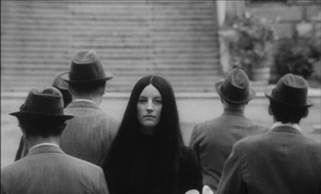 Black and white image of a woman with long black hair is standing in the center of a group of men. The men have their backs turned to the camera. Behind them is an old concrete staircase.