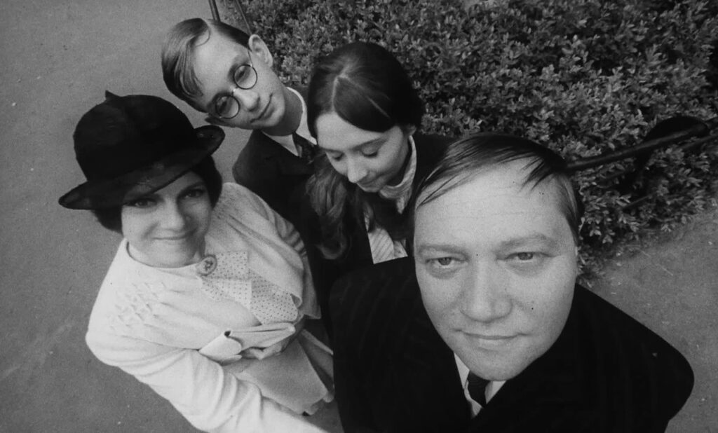 High angle black and white shot of a family of four outside in a park. The father and the mother are looking directly into the camera, their children (son and daughter) are looking down. They are dressed in fine clothing, suits and blazers, and there is a gated off bush behind them.