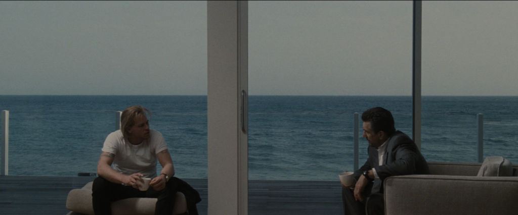 Two men, the blond-haired Chris Shiherlis (played by Val Kilmer) and the brown-grey-haired Neil McCauley (played by Robert DeNiro), sit in off-white chairs in a large living room in a beachfront house. Chris is wearing a plain white T-shirt and Neil is wearing a blue-gray suit. Both of them hold coffee cups as they sit facing each other. Behind them, we can see the waves of the ocean just beyond the sliding glass doors