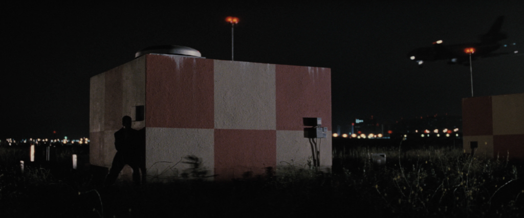 A shadowy silhouette of a man leans against a red-and-white checkered block structure in the middle of an airport runway. Other than the block and the lights atop the structure, as well as the ones dotting the skyline, it is too dark at night to make out much other detail.