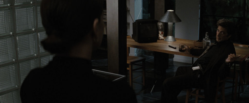 Vincent Hanna (played by Al Pacino), a middle-aged man with dark brown hair, sits at a dining table, holding a glass of hard liquor. He is looking at a woman, whose back faces the camera. There is a small television and lamp on the dining table.