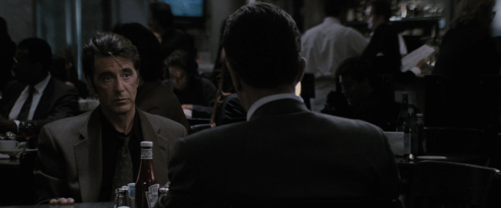 Vincent Hanna (played by Al Pacino), a middle-aged man with dark brown hair and a gray-and-black suit, sits at a diner table with sugar packets and ketchup, opposite another man, who we can only see the back of.