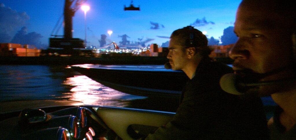 I’m becoming this James “Sonny” Crockett (Colin Farrell) and Ricardo “Rico” Tubbs (Jamie Foxx) in a speedboat on water at sunrise, staring pensively ahead while driving past shipping containers.