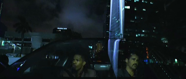  James “Sonny” Crockett (Colin Farrell) and Ricardo “Rico” Tubbs (Jamie Foxx) driving through downtown Miami at night, lit up skyscrapers reflected in the windshield of their vehicle.