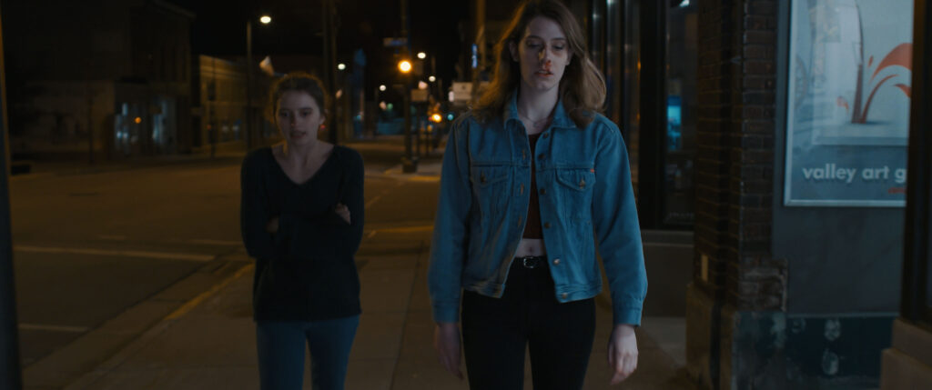 Medium Wide of two young women walking down an empty small town, midwestern, downtown street at night.
