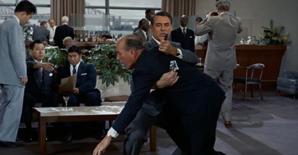 Cary Grant as Roger Thornhill in North By Northwest holding up a man with a knife in his back. Thornhill is holding the man up with his right arm and gripping the knife with his left hand. Several men are noticing behind him with one pointing. 