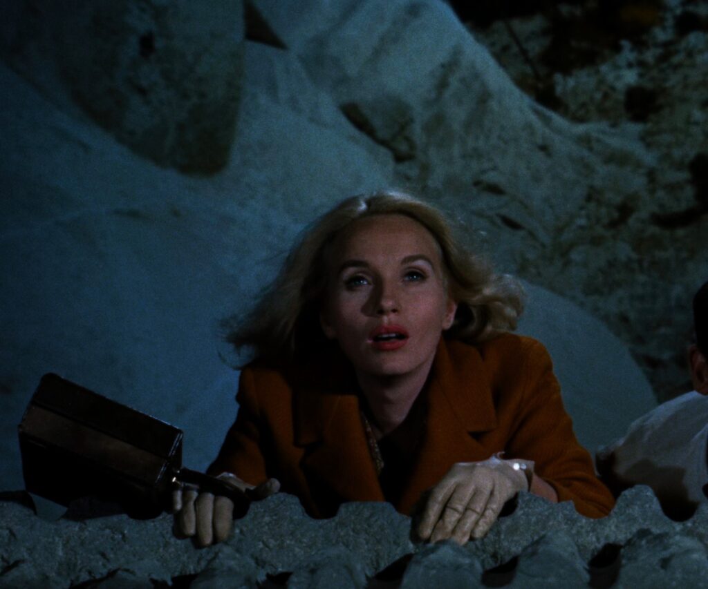 Screenshot 5: Close up image of Eva Marie Saint hanging bent armed on the ledge of Mt Rushmore. Her purse hangs out to the side with a painted backdrop of Mt Rushmore behind her.
