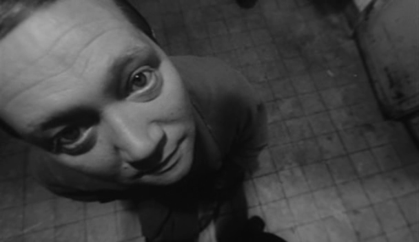 A high-angle, black and white, fish-eye close up of a man in a suit. his eyes are slightly bulging. the floor below him is dilapidated tile.