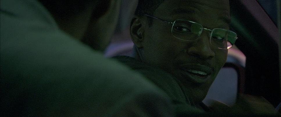 Close up over the shoulder shot of Jamie Foxx. He's smiling slyly, wearing glasses, and is in the front seat of a taxi.